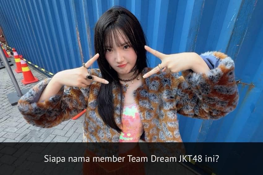 Seorang anggota Team Dream JKT48 berpose dengan gaya tangan membentuk simbol di depan kontainer biru di area luar ruangan.
