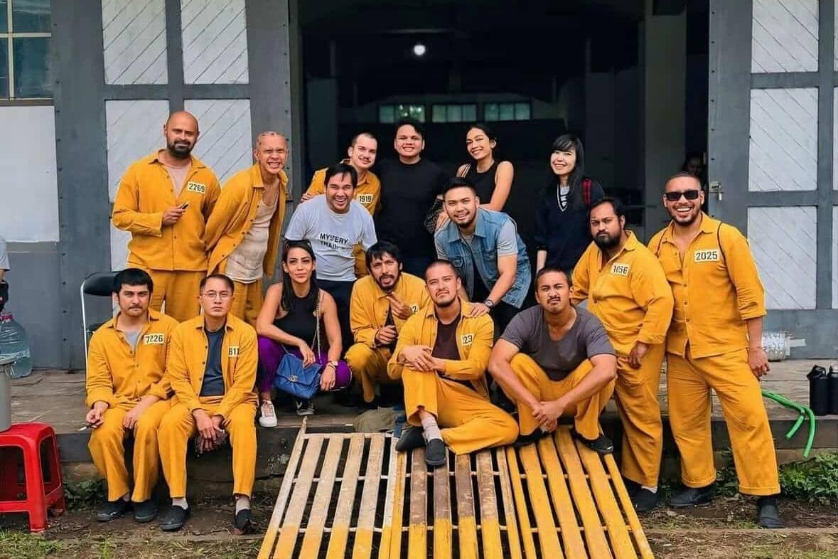 Sekelompok orang berpose bersama di depan bangunan, sebagian mengenakan seragam tahanan berwarna oranye dengan nomor identitas.