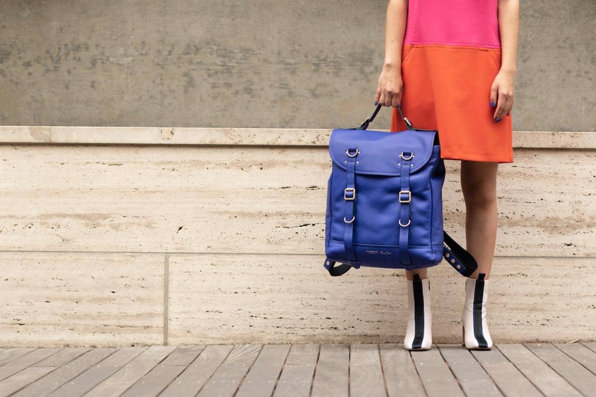 Seorang perempuan mengenakan gaun merah muda dan oranye memegang tas ransel kulit berwarna biru dengan detail gesper logam.