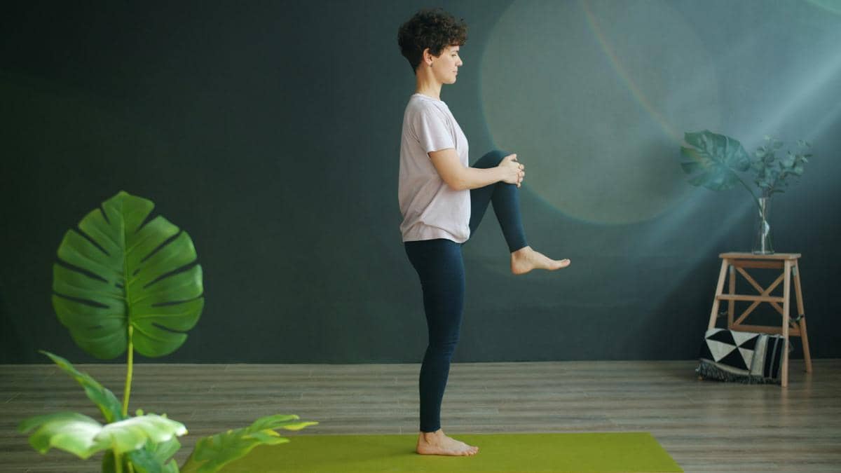 Seseorang berdiri di atas satu kaki di atas matras yoga hijau sambil memegang lutut, melakukan latihan keseimbangan di ruangan minimalis.