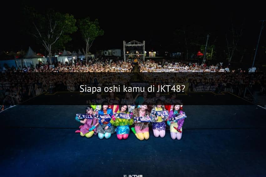 Anggota grup idola JKT48 berpose di atas panggung dengan kostum warna-warni di depan penonton yang memenuhi area konser malam hari.