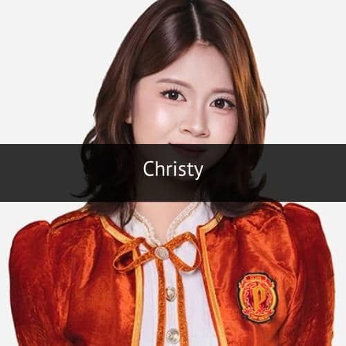 Seorang anggota JKT48 mengenakan seragam berwarna oranye dengan detail pita dan emblem di dada, berpose dengan latar polos.