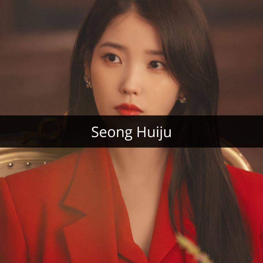 Perempuan berambut panjang mengenakan blazer merah duduk di kursi dengan ekspresi serius di ruangan bercahaya hangat.