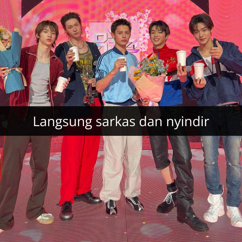 Lima pria muda berpose di atas panggung dengan latar merah muda sambil memegang bunga dan cangkir kertas setelah penampilan.