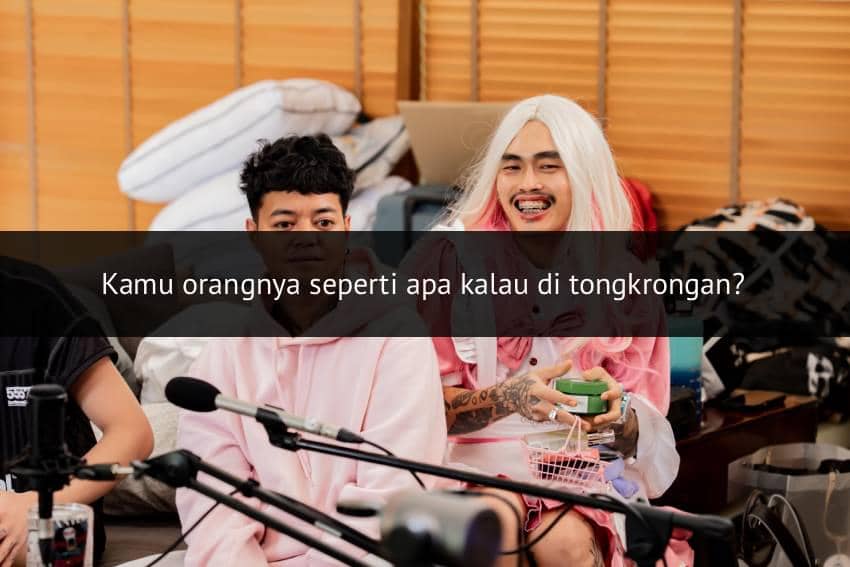 Dua pria duduk bersama di ruangan dengan peralatan rekaman, salah satunya mengenakan wig pirang panjang dan pakaian berwarna pink.