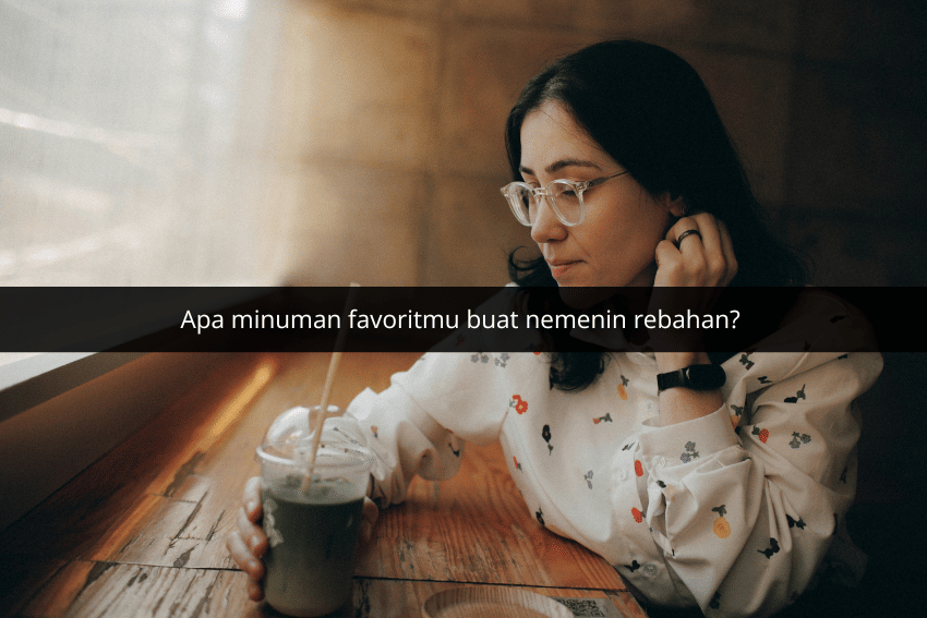 Seorang wanita berkacamata duduk di dekat jendela sambil memegang minuman dingin dengan sedotan di tangan kanan.