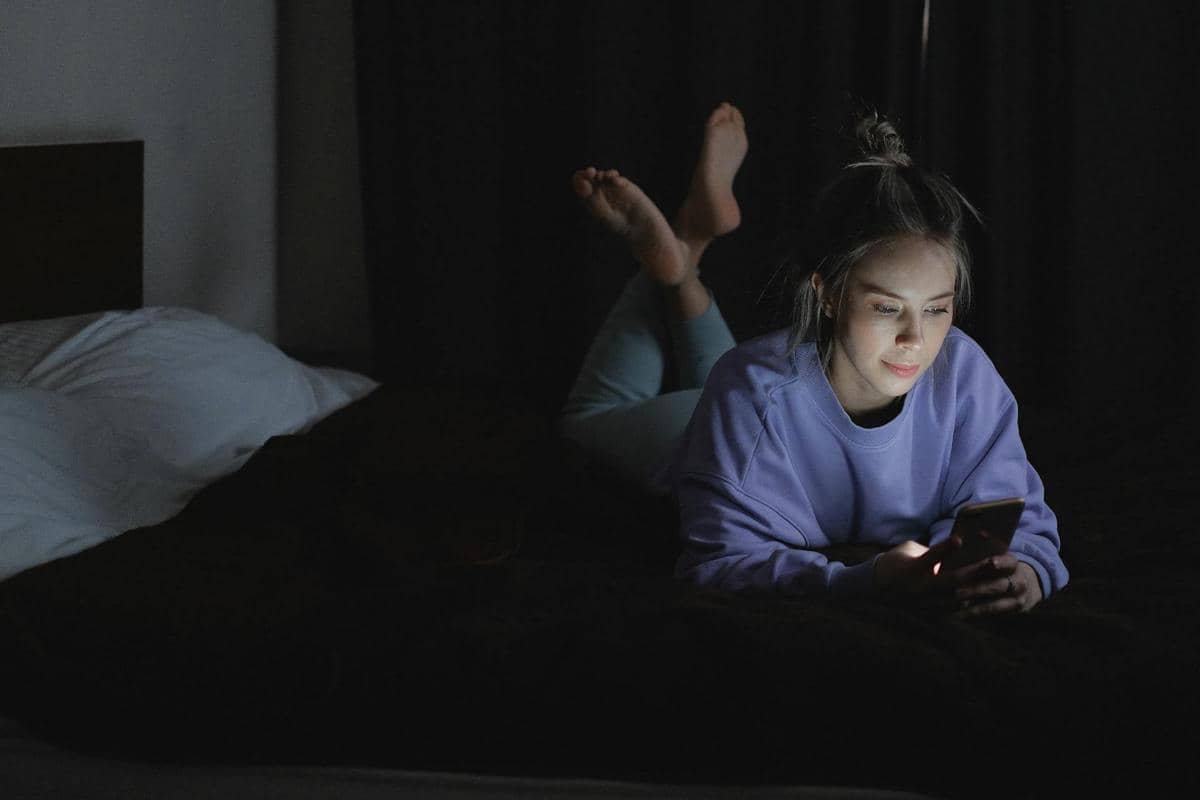 Perempuan berbaring di tempat tidur sambil menggunakan ponsel pada malam hari.