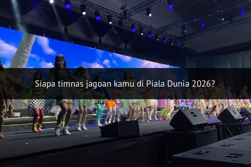 Para anggota grup idola tampil menari di atas panggung dengan pencahayaan warna-warni dan latar layar besar bergambar langit biru.