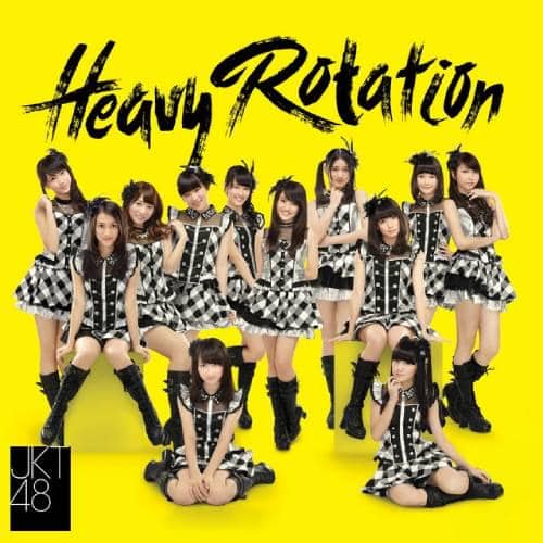 Grup idola JKT48 mengenakan kostum hitam putih bermotif kotak-kotak berpose di depan latar kuning dengan tulisan Heavy Rotation.