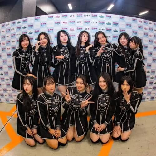 Sebelas anggota grup idola perempuan berpose bersama mengenakan kostum hitam dengan garis putih di depan latar bertuliskan berbagai logo sponsor.
