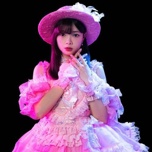 Seorang anggota JKT48 mengenakan gaun dan topi berwarna merah muda dengan pencahayaan ungu, berpose di latar belakang hitam.