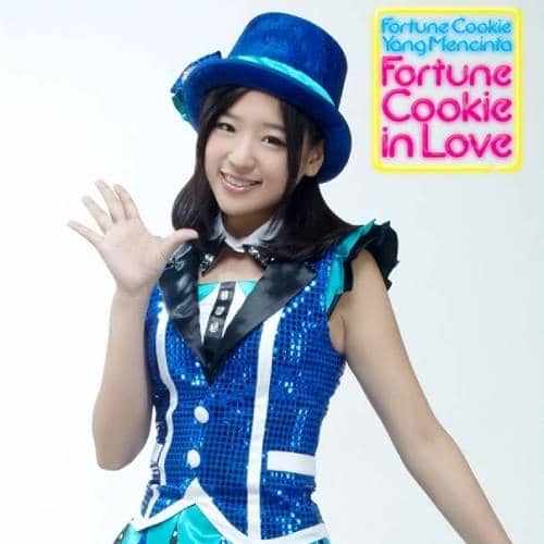 Seorang anggota grup idola JKT48 mengenakan kostum biru berkilau dan topi biru, tersenyum sambil melambaikan tangan di depan latar putih.