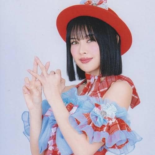 Seorang anggota JKT48 mengenakan kostum panggung berwarna merah dan biru dengan topi merah, berpose dengan tangan membentuk simbol huruf.