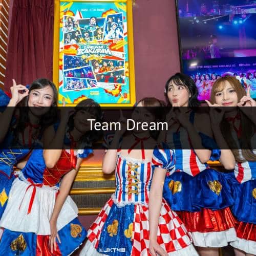 Beberapa anggota grup idola JKT48 mengenakan kostum warna merah, biru, dan putih berpose ceria di depan poster bertema Team Dream.
