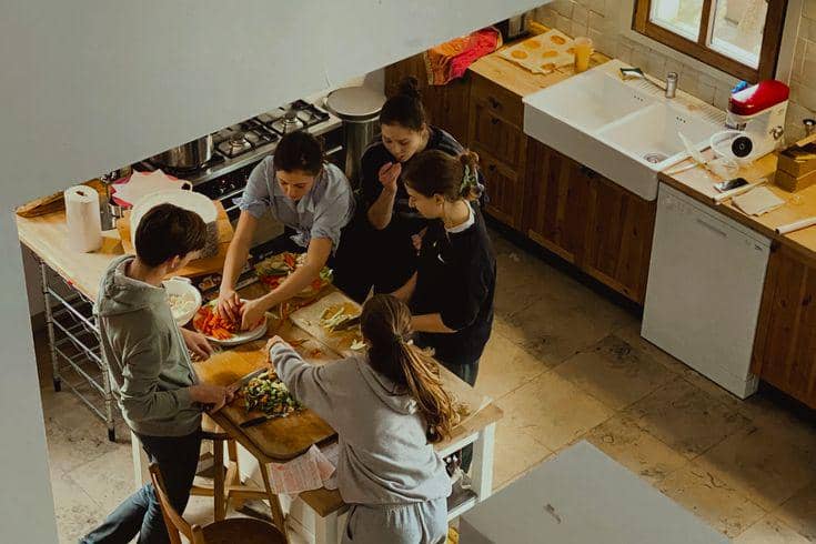 Sudut pandang tinggi menampilkan sekelompok anak muda yang sedang berkumpul di sekitar meja pulau kayu di dapur untuk memotong sayuran dan menyiapkan makanan bersama di dalam rumah berasitektur kayu hangat.
