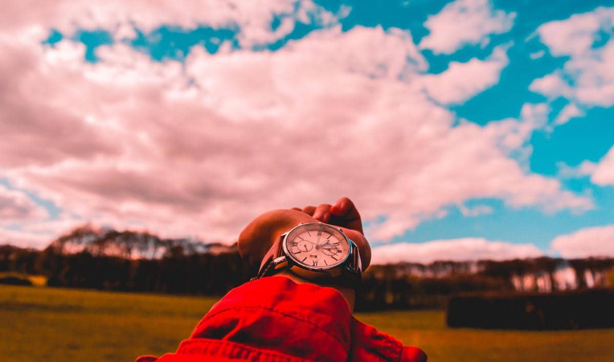 Tangan mengenakan jam tangan analog dengan latar langit berawan dan pemandangan padang rumput yang luas di siang hari.