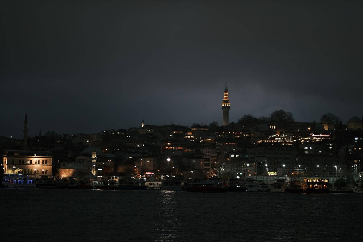 Pemandangan kota di tepi laut pada malam hari dengan gedung-gedung bercahaya dan menara tinggi yang diterangi lampu kuning.