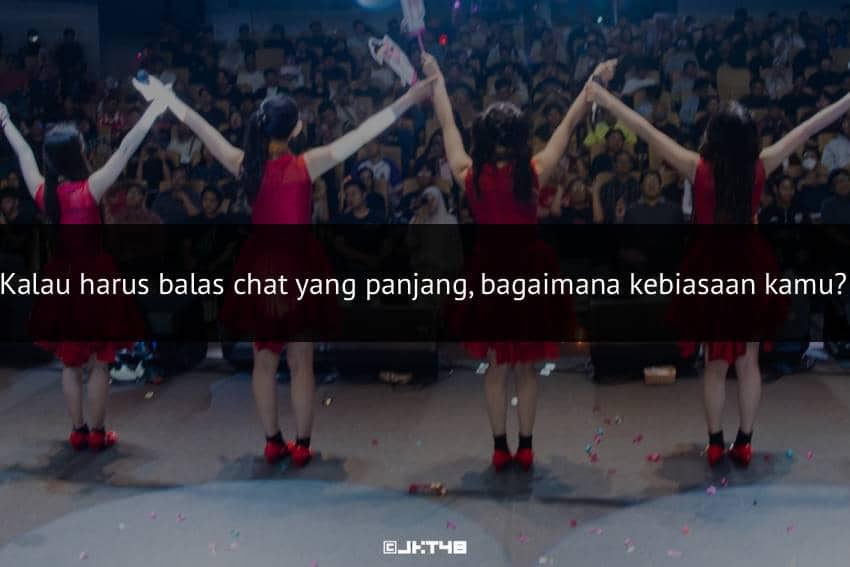 Empat anggota grup idola mengenakan gaun merah berdiri di atas panggung sambil bergandengan tangan menghadap penonton yang memenuhi ruangan.