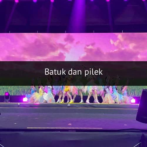 Grup idola perempuan tampil di atas panggung dengan kostum warna-warni di bawah pencahayaan ungu dan latar layar besar bernuansa senja.