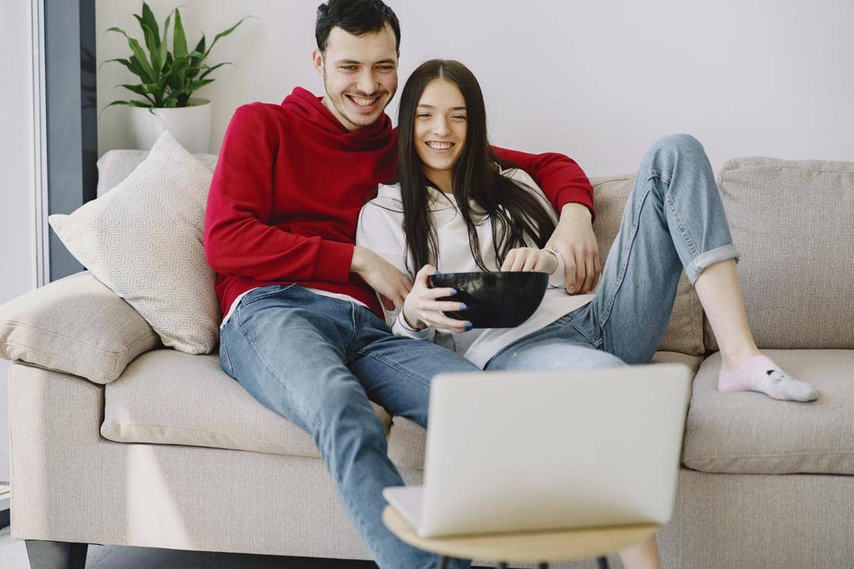 Pasangan pria dan wanita tersenyum bahagia saat bersantai menonton film melalui layar laptop di atas sofa.