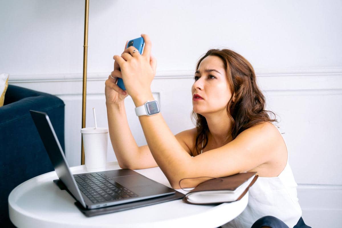 Wanita duduk di meja kerja memegang HP dengan ekspresi bosan dan laptop terbuka.