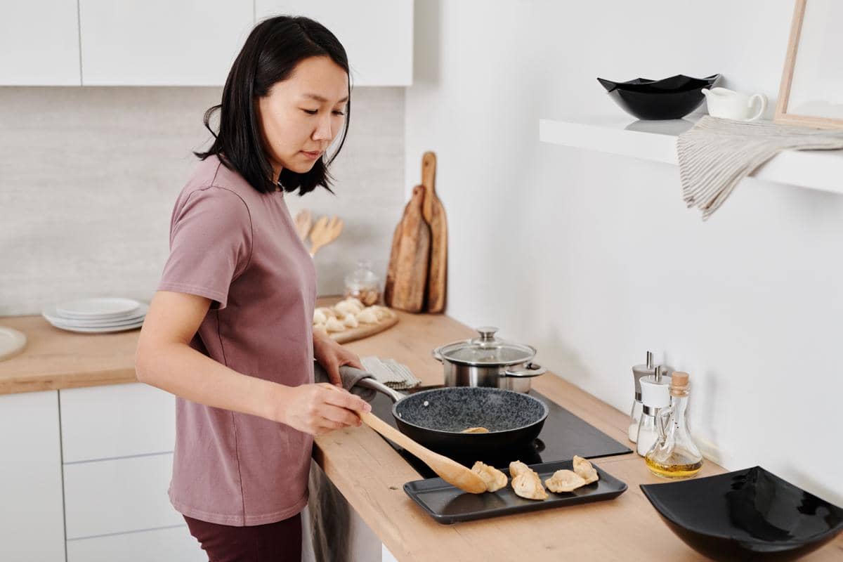 Seorang perempuan sedang memasak makanan di atas wajan menggunakan kompor dapur modern.